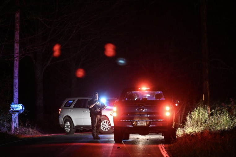 An armed police officer stands on the road with emergency vehicles